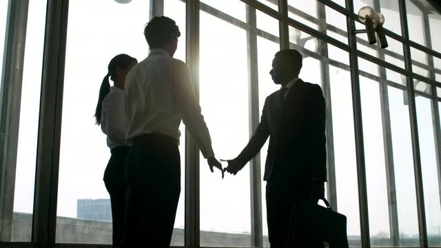 Medium shot of black businessman in suit walking towards male and female business partners and exchanging handshake in greeting before Panoramic windows on sunny day