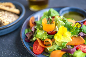Close up view of vegetable salad with fresh tomatoes, flowers, carrot slices and different summer greens, served with olive oil on blue plate, dark background. Healthy, vegan, diet, raw food concept.