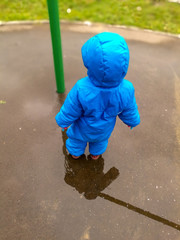 A little boy in a blue jumpsuit stands in the middle of a puddle next to a post on the Playground