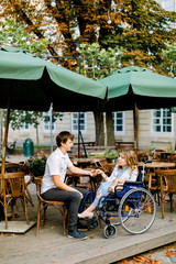 Young pretty woman in wheelchair spending time together with male boyfriend, holding hands, at outdoor cafe in the old city center