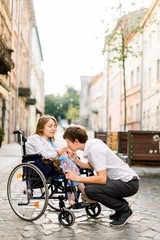 Handsome happy man smiling and holding hand of his beautiful beloved blond handicapped woman in wheelchair while walking together on the street of old city