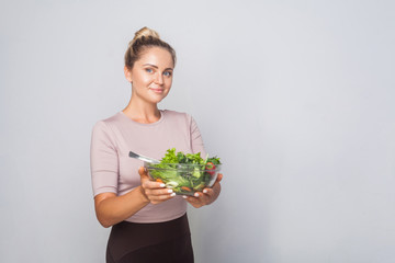 Portrait of cheerful fit woman with hair bun holding bawl of green vegetable salad and looking at camera, vegetarian diet, healthy food, empty copy space for advertising. studio shot, grey background