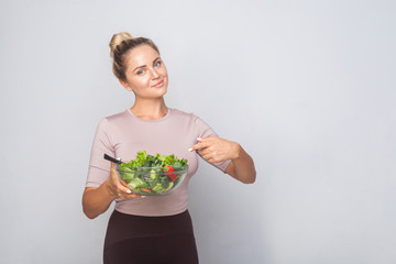 Portrait of satisfied fit woman with hair bun pointing at bawl of vegetable salad and looking at camera, vegetarian diet, healthy food, empty copy space for advertising. studio shot, grey background