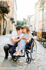 Young man hugs his pretty woman on the wheelchair, while walking together on the street of city. Couple in love in wheelchair embracing and touching foreheads with closed eyes, outdoors in the city
