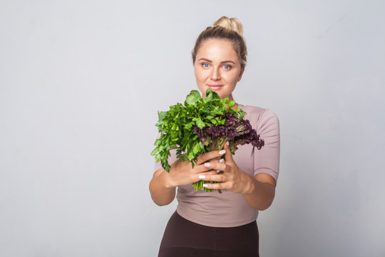 Young positive girl giving bunch of fresh cooking herbs to camera and smiling, leafy green vegetables, holding parsley sorrel lettuce, healthy nutrition, organic food. studio shot, grey background