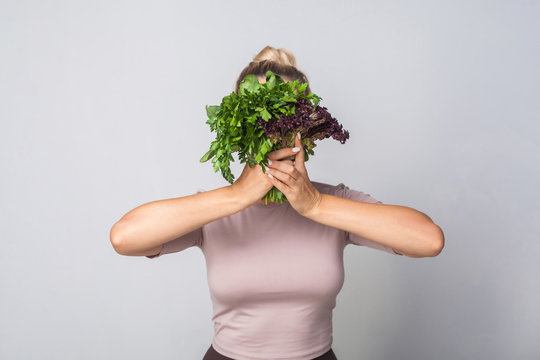 Young Woman Hiding Face Behind Bunch Of Herbs, Leafy Green Vegetables, Holding Parsley Sorrel Lettuce And Smiling Looking At Camera, Healthy Nutrition, Organic Food. Studio Shot, Grey Background