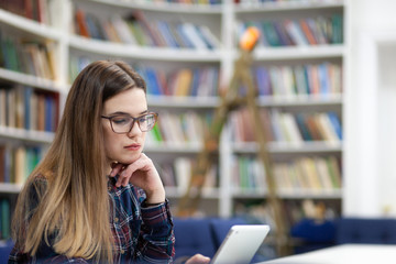 Girl in glasses working with tablet sitting in library. Student prefers to work with a tablet than books