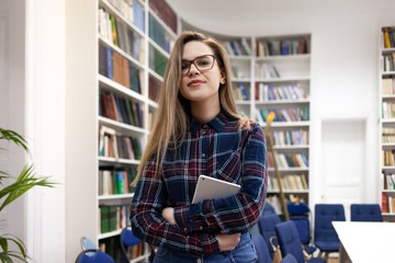 Beautiful young female student is standing in a college library room against the background of shelves with books holding a tablet in her hands. Portrait of a girl in the library