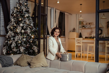 Portrait of a stylish young woman in a white jacket who holds a round box with a gift in her hands. She is in a beautiful room with a Christmas tree.