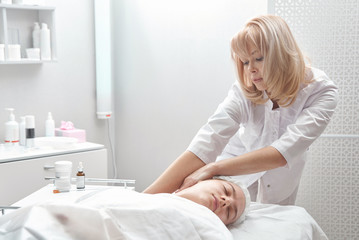 Obraz na płótnie Canvas Professional beautician gives young woman manual massage of face and cervical collar zone in white beauty parlor. Сosmetology, face care procedure