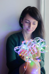young european woman sitting on windowsill and holding light colorful garland, vertical stock photo image