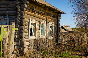 An old abandoned log house in the Russian Outback with slanted Windows and shutters and a dilapidated roof.