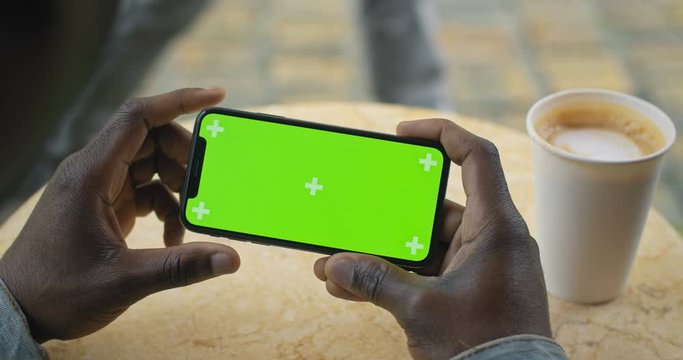 Hands of Afro American Man Sits at Table with Paper Coffee Cup on it Holding Smartphone Horizontal with Green Screen, Scrolling, Touching Screen.