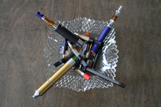 Closeup of assorted vape pens and cartridges in a glass ashtray.