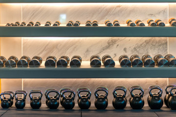 Wide range of choices. Kettlebells weights and dumbbells lying in a row on shelves in stylish workout gym. Marble background. Sport, healthy lifestyle concept. Horizontal shot.