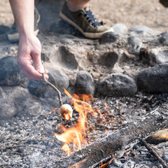 Tasty, sweet marshmallow on a crooked twig is held by a male hand over a bonfire. Cooking dessert on campfire.