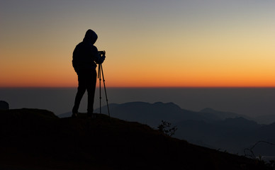 silhouette of Travel photographer standing with a camera mounted on a tripod and shooting a time lapse of the sunrise/sunset. man wearing his hood enjoying the mountain view /valley view
