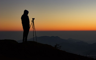silhouette of Travel photographer standing with a camera mounted on a tripod and shooting a time lapse of the sunrise/sunset. man wearing his hood enjoying the mountain view /valley view