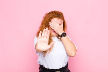 young red head woman covering face with hand and putting other hand up front to stop camera,...