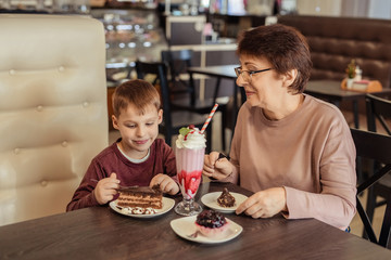 A happy grandmother with glasses and her grandson spend 7 years together in a cafe. they eat sweet cakes and milkshakes.