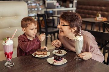 A happy grandmother with glasses and her grandson spend 7 years together in a cafe. they eat sweet cakes and milkshakes.