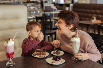 A happy grandmother with glasses and her grandson spend 7 years together in a cafe. they eat sweet cakes and milkshakes.