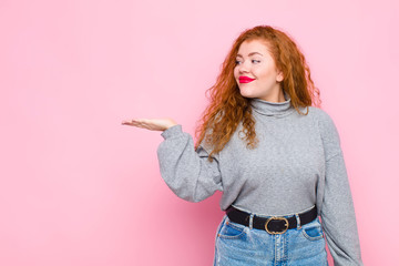 young red head woman feeling happy and smiling casually, looking to an object or concept held on...