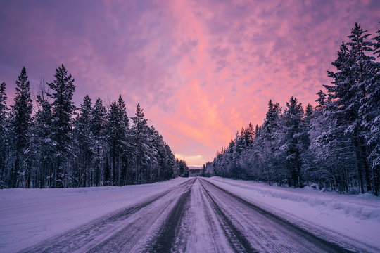 Remote winter road through snow covered forest trees against dramatic purple pink sky, Lapland, Finland