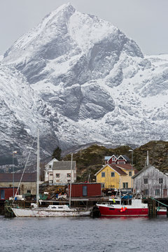 Fishing village boats at waterfront below snowy, rugged mountains, Sund, Lofoten, Norway
