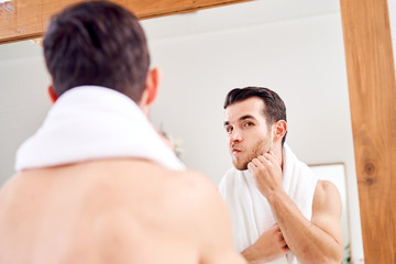 Fototapeta premium Unshaven male with white towel on his neck standing near mirror