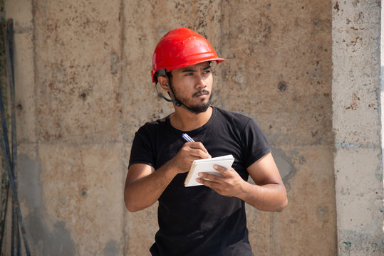 Black bearded engineering wear red hardhat and black working suit recording work and make determined face in construction site