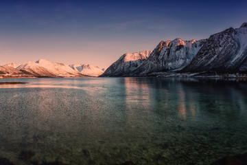 Amazing winter landscape with rocky snowy mountains in sunset light, blue sky and reflection in water, Lofoten Islands, Norway. Outdoor travel background