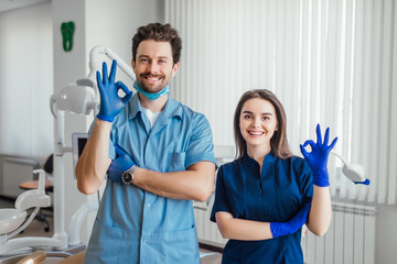 Photo of smiling dentist standing with arms crossed with her colleague, showing okay sign.