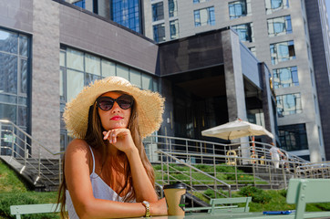 beautiful slender tanned woman in a straw hat and a white sundress sits at a table on a summer terrace of a cafe and drinks coffee.