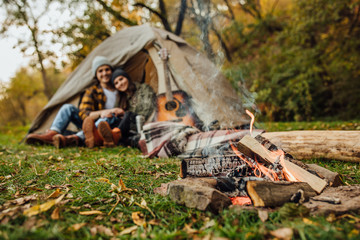 Focus on fire near tent. Young loved couple of tourists have a date in the forest. Attractive young woman and handsome man are spending time together on nature. Hugging lovers near campfire.