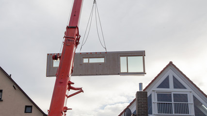 Completed component with a large window of a prefabricated house on a mobile crane boom against the sky between two houses