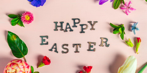 Happy Easter letters with different spring flowers around them, flat lay, pink background, banner image