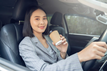 Asian female driver smiling and drinking coffee in the car, Beautiful girl  holding an eco paper coffee cup,looking to camera while driving her car, happy life transport in city