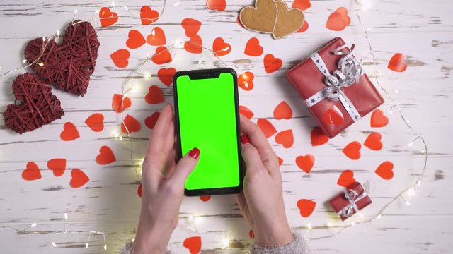 Valentine's Day, a woman uses a smartphone with a green screen on a desk decorated with hearts, close-up top view.