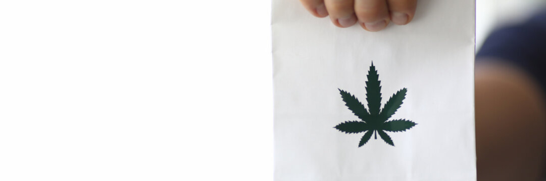 Focus On Male Hand Holding White Small Paper Bag With Green Cannabis Symbol On It. Healthcare And Medicines Concept. Copy Space In Left Side. Blurred Background