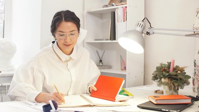Young professional designer woman in white formal shirt sit at table writing in a notebook, wearing eyeglasses, sit in office
