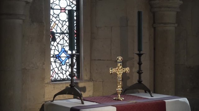 Close-up view of the large golden cross standing on the table between two black candles against the window with stained glass. Action. Religion and faith