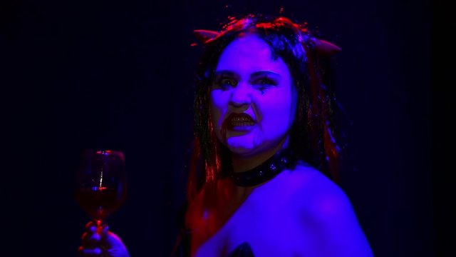 Woman devil with an ominous smile on her face drinks blood from a glass like a wine face in blood.