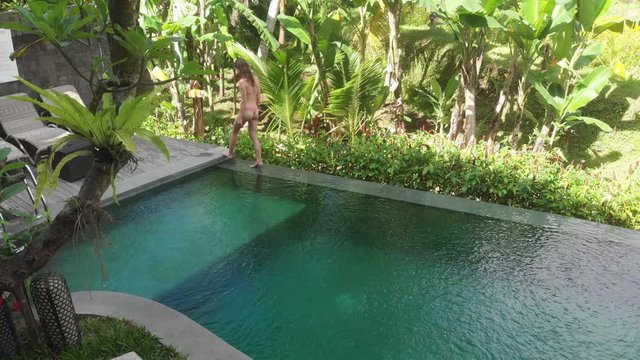 Aerial View of Infinity pool on villa with tropic garden. Woman walking on edge of pool and enjoy jungle view wearing beige bikini and hat. Vacation concept