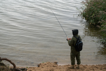 Fisherman standing on sandy shore of lake near a thicket of green reeds, holding fishing rod with tackle thrown into water dressed in  green protective suit with hood with a backpack on his shoulders