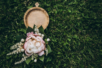 Round wooden box with tender flowers, greenery, roses and golden wedding rings, lying on a fresh green grass.