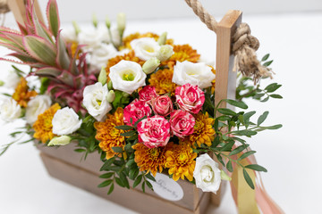 Unusual expressive floral arrangement of fresh flowers with satin multi-colored ribbons (colors: white, orange, red, green) in a gift wooden box with a pen made of rope on a white background