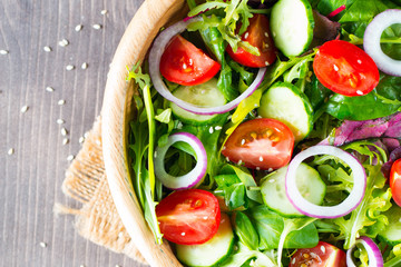 Fresh healthy salad with delicious ruccola, spinach, cabbage, arugula, feta cheese, red onion, cucumber, sesame seeds and cherry tomato on wooden background. Healthy and diet food concept.