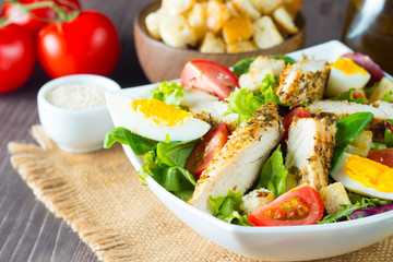 Fresh Caesar salad with delicious chicken breast, ruccola, spinach, cabbage, arugula, egg, parmesan and cherry tomato on wooden background. Oil, salt and pepper. Healthy and diet food concept.