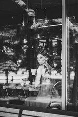 Fototapeta na wymiar Beautiful Serious European brunette girl in an evening red dress sitting at the window with a cup of coffee at the cafe. Look at the camera through the window. black and white photo